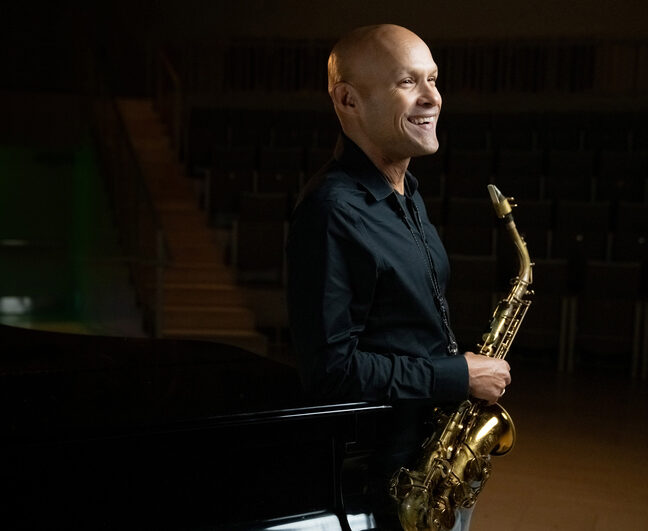 Faculty member holding saxophone while leaning against piano.